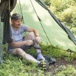 Person sitzt im Schatten eines grünen Trekkingzelts im Naturpark Neckartal-Odenwald und bindet ihre Wanderschuhe, umgeben von Grünpflanzen.