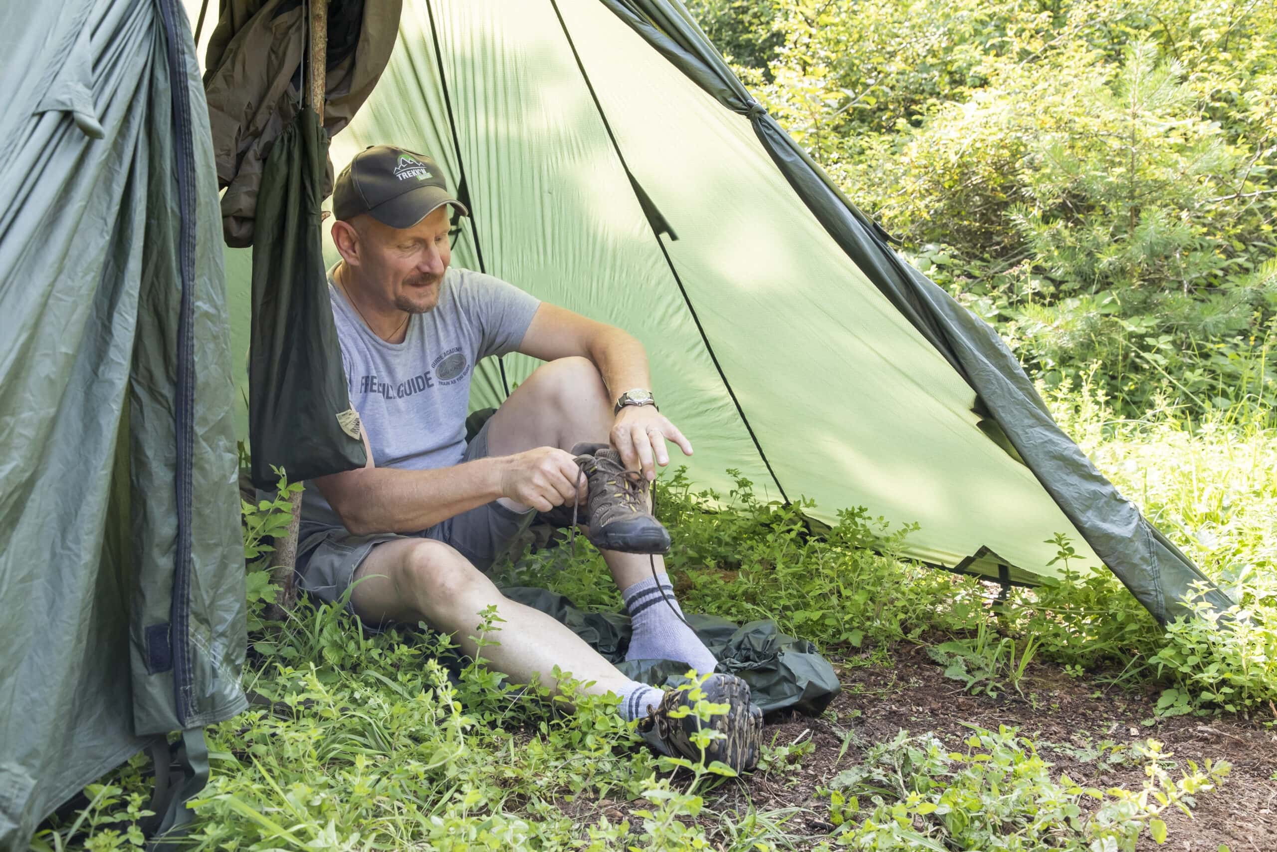 Person sitzt im Schatten eines grünen Trekkingzelts im Naturpark Neckartal-Odenwald und bindet ihre Wanderschuhe, umgeben von Grünpflanzen.