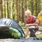 Drei Personen richten im Wald des Naturparks Neckartal-Odenwald ein grünes Trekkingzelt auf einem kiesigen Untergrund auf.