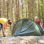 Zwei Personen montieren im schattigen Wald des Naturparks Neckartal-Odenwald ein grünes Trekkingzelt auf einem kiesigen Untergrund.