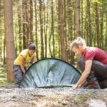 Zwei Personen richten im Naturpark Neckartal-Odenwald ein grünes Trekkingzelt auf einem moosigen Waldboden zwischen dichtem Fichtenwald auf.