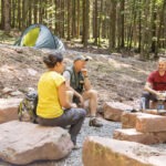 Drei Personen sitzen auf Felsen in einem Waldcamp im Naturpark Neckartal-Odenwald, während im Hintergrund ein Zelt und eine einfache Camp-Kochstelle mit Gaskocher zu sehen sind.