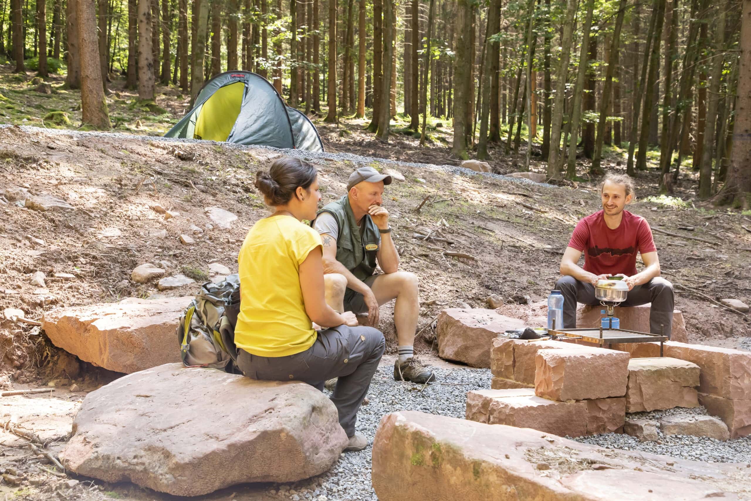 Drei Personen sitzen auf Felsen in einem Waldcamp im Naturpark Neckartal-Odenwald, während im Hintergrund ein Zelt und eine einfache Camp-Kochstelle mit Gaskocher zu sehen sind.