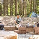Zwei Personen sitzen auf einer Steinbank in einem Waldcamp im Naturpark Neckartal-Odenwald, während im Hintergrund ein Zelt und eine einfache Feuerstelle mit Kochutensilien zu sehen sind.