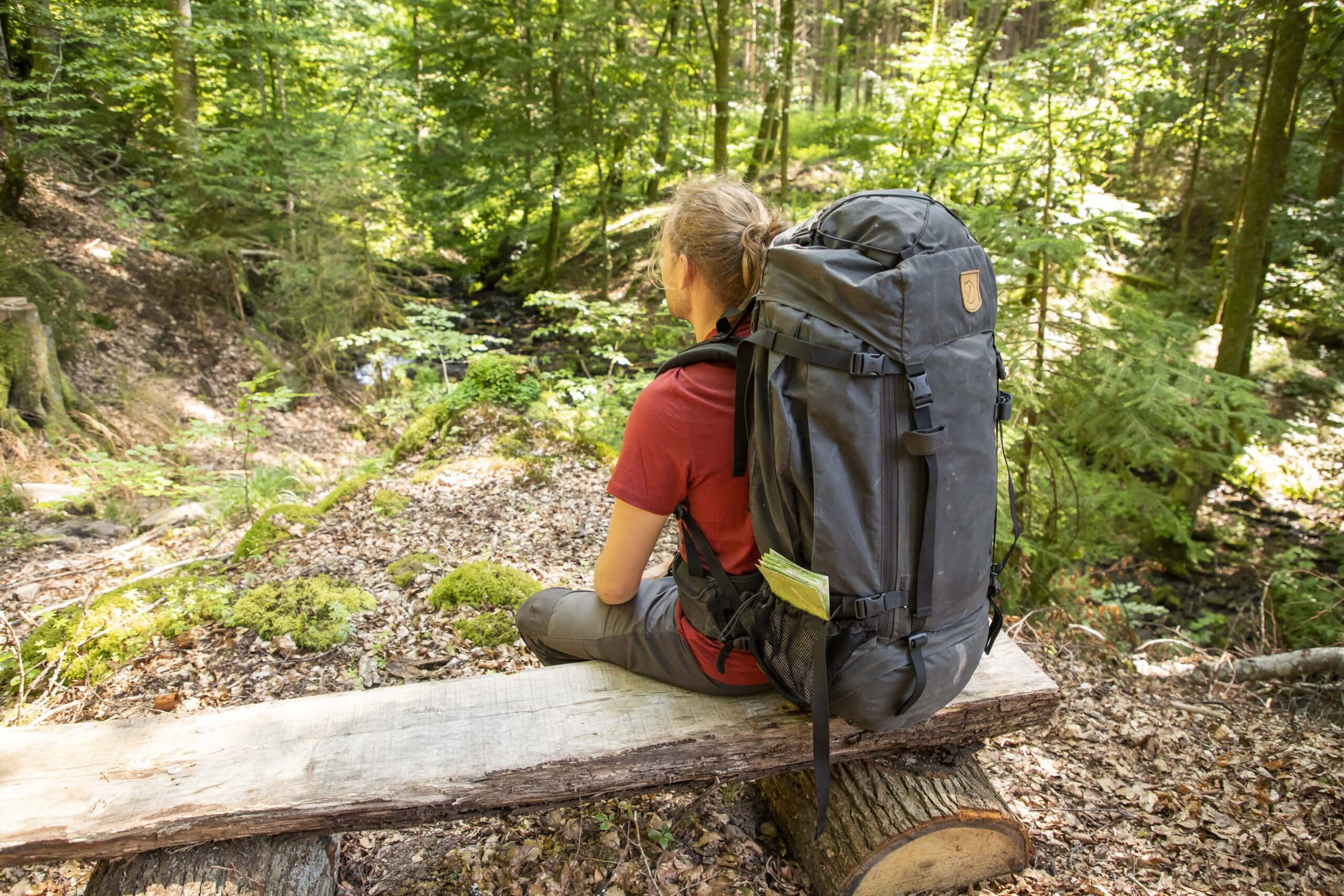 Person mit großem Trekkingrucksack sitzt auf einem Baumstumpf im schattigen Wald des Naturparks Neckartal-Odenwald und pausiert.