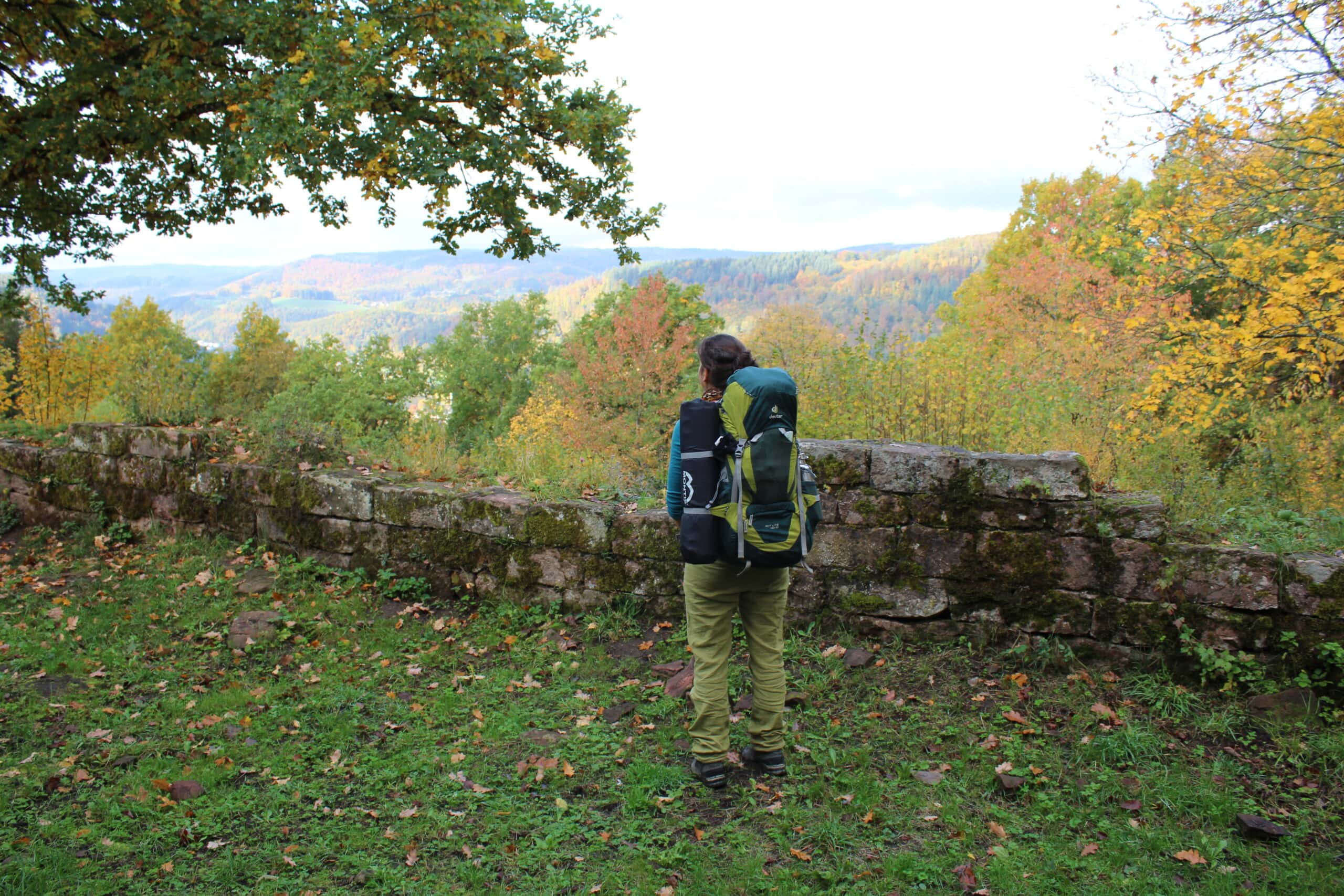 Person mit Rucksack lehnt an einer alten Steinmauer und betrachtet die herbstliche Landschaft mit buntem Laub im Naturpark Neckartal-Odenwald.