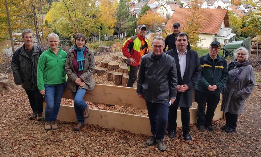 Gruppe von acht Personen vor einem Hirschkäfermeiler aus Holz und Laub, umgeben von herbstlichem Wald und Häusern im Hintergrund.