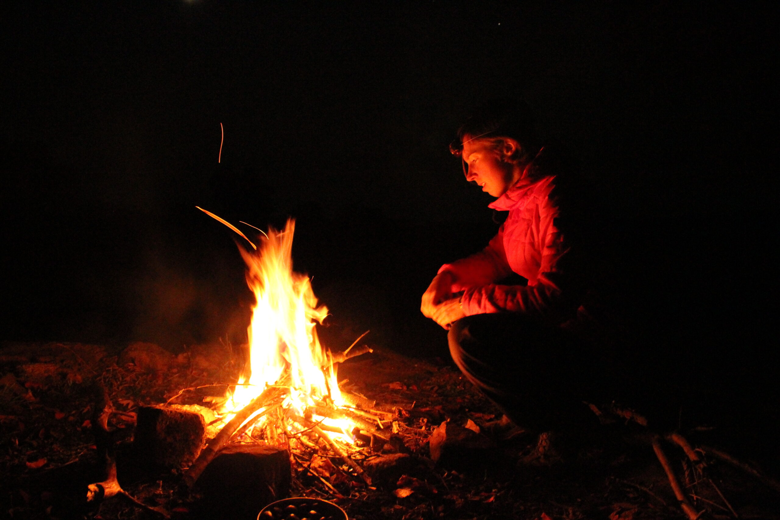 Person sitzt abends am Lagerfeuer im Freien, umgeben von Dunkelheit und Flammenlicht, typisch für eine Pause beim Trekking.