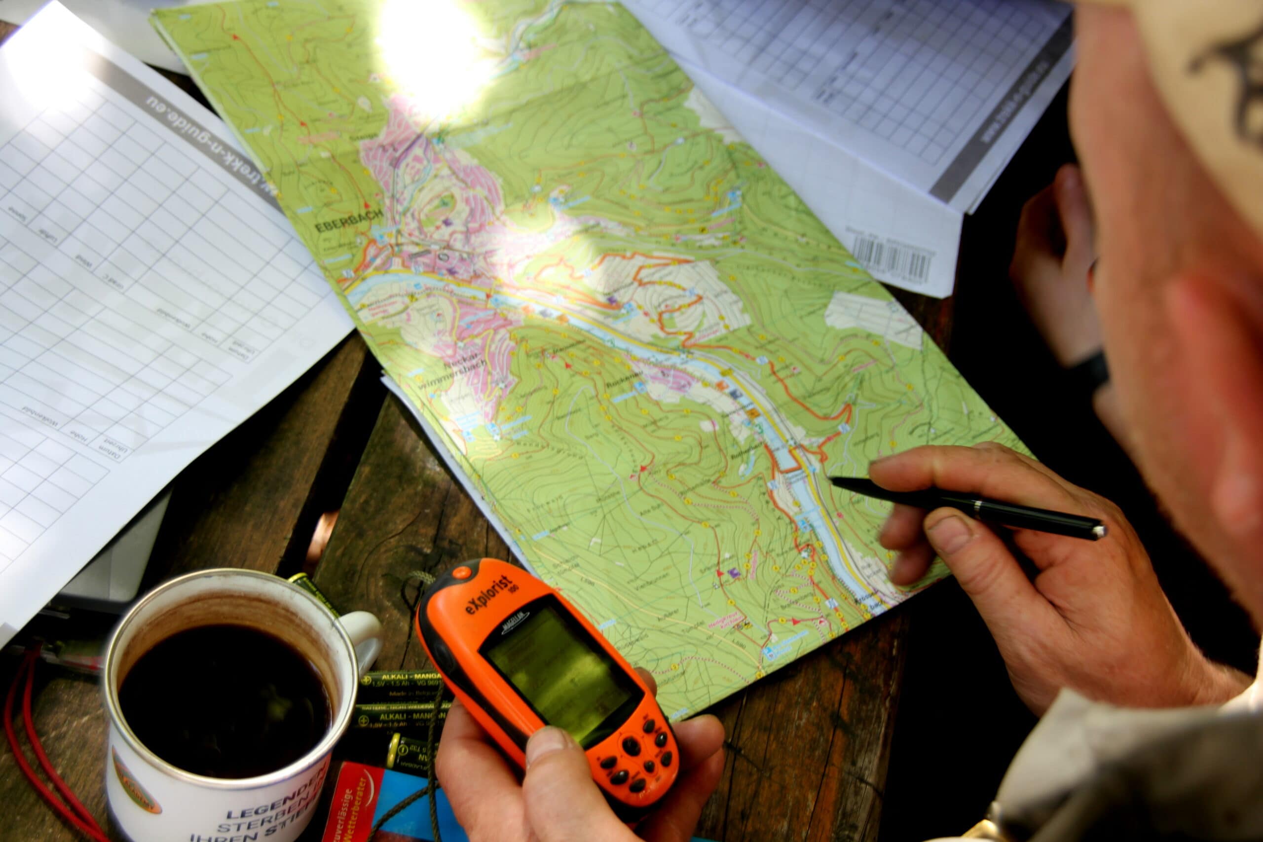 Person plant Wanderroute im Naturpark Neckartal-Odenwald mit GPS-Gerät und detaillierter Wanderkarte, Tasse mit Heißgetränk daneben.