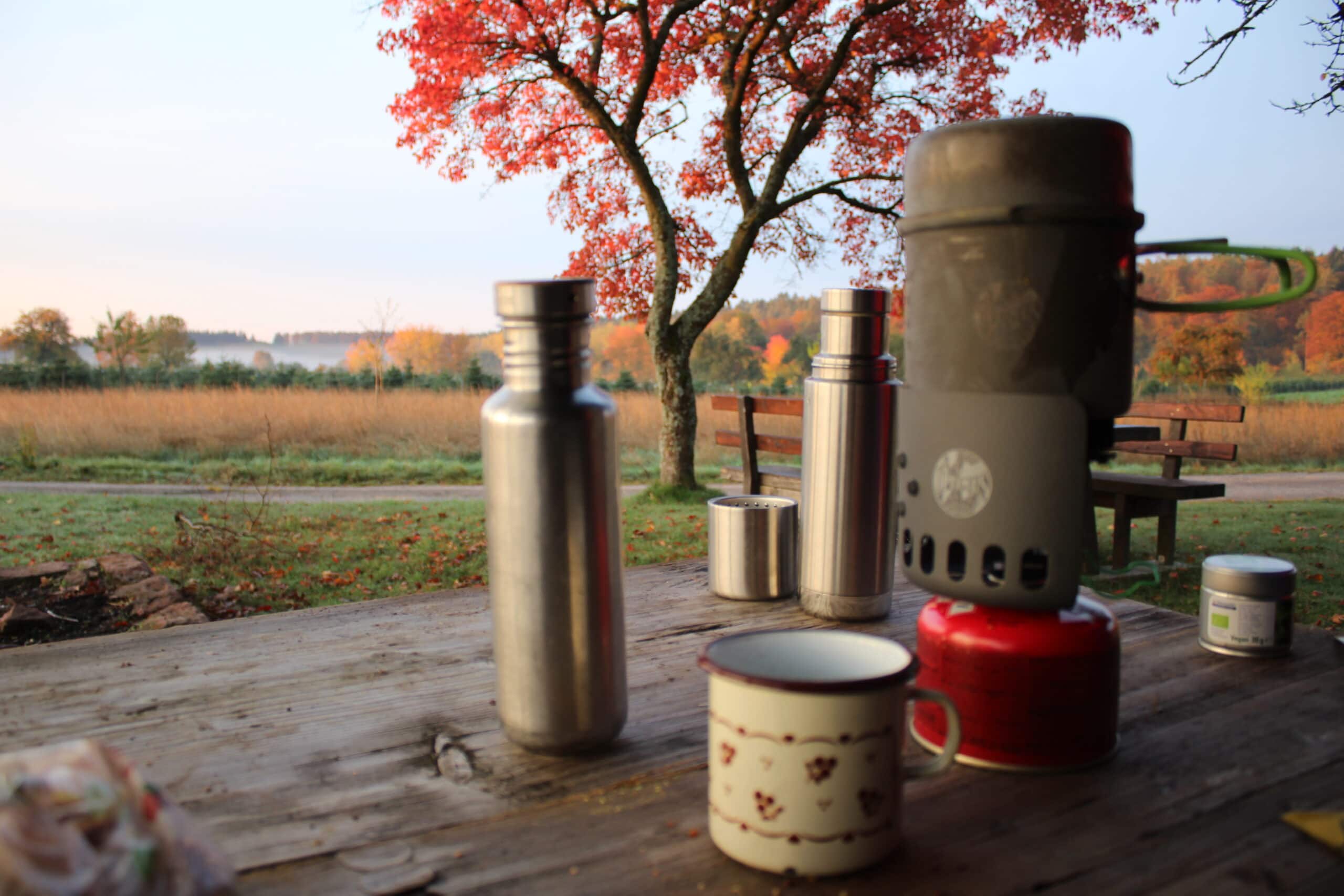 Campingausrüstung auf Holztisch: Gaskocher, Thermoskanne, Metallbehälter, Tasse und Zutatenbehälter vor herbstlich gefärbtem Baum und Waldweg im Naturpark.