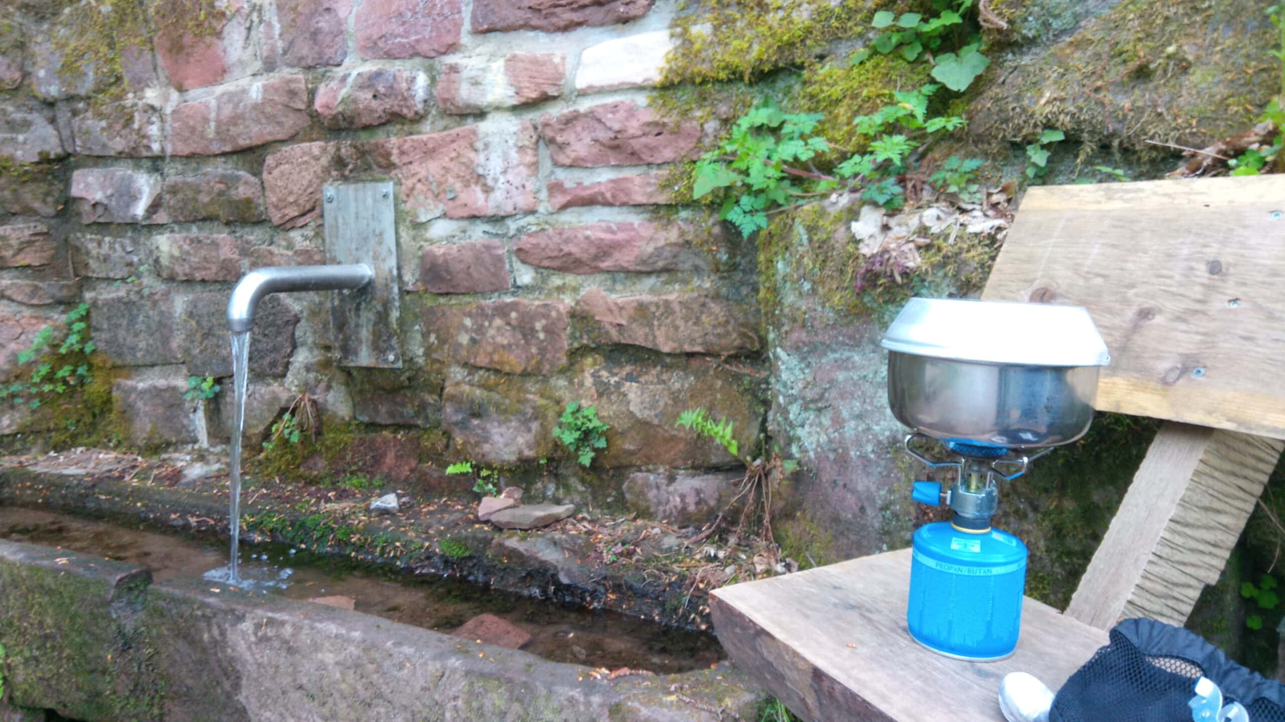 Wasserspender mit fließendem Wasser neben moosbewachsenen Steinmauer, daneben Campingkocher auf Holztisch in waldiger Umgebung.