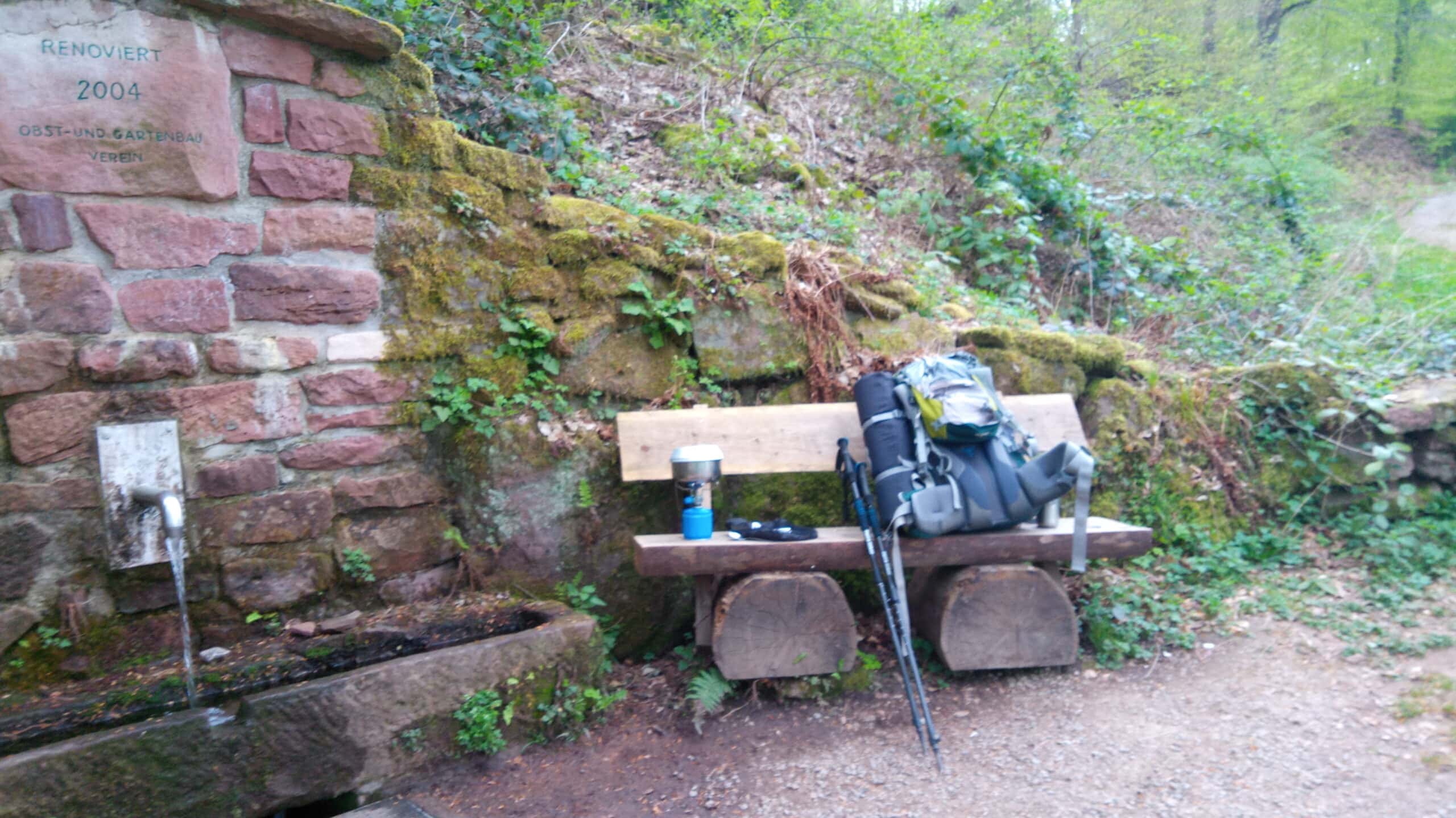 Holzbank mit Rucksack, Trekkingstöcken und Kochgeschirr an einer moosbewachsenen Steinmauer im Wald, typisch für eine Rastpause im Naturpark Neckartal-Odenwald.