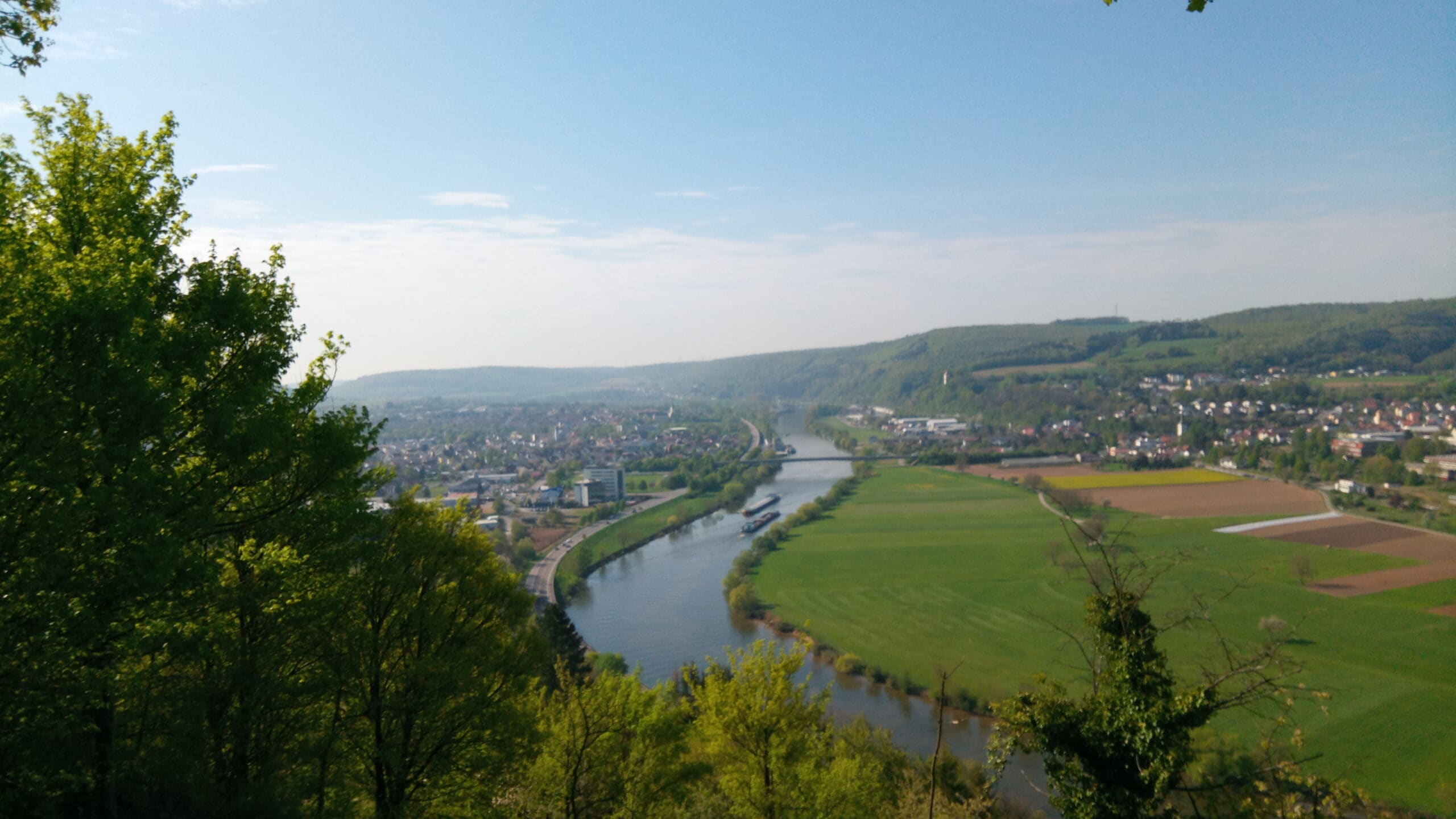 Weitläufige Neckartal-Landschaft im Naturpark: Fluss windet sich durch grüne Wiesen und Wälder, umgeben von sanften Hügeln und einem Dorf im Hintergrund.