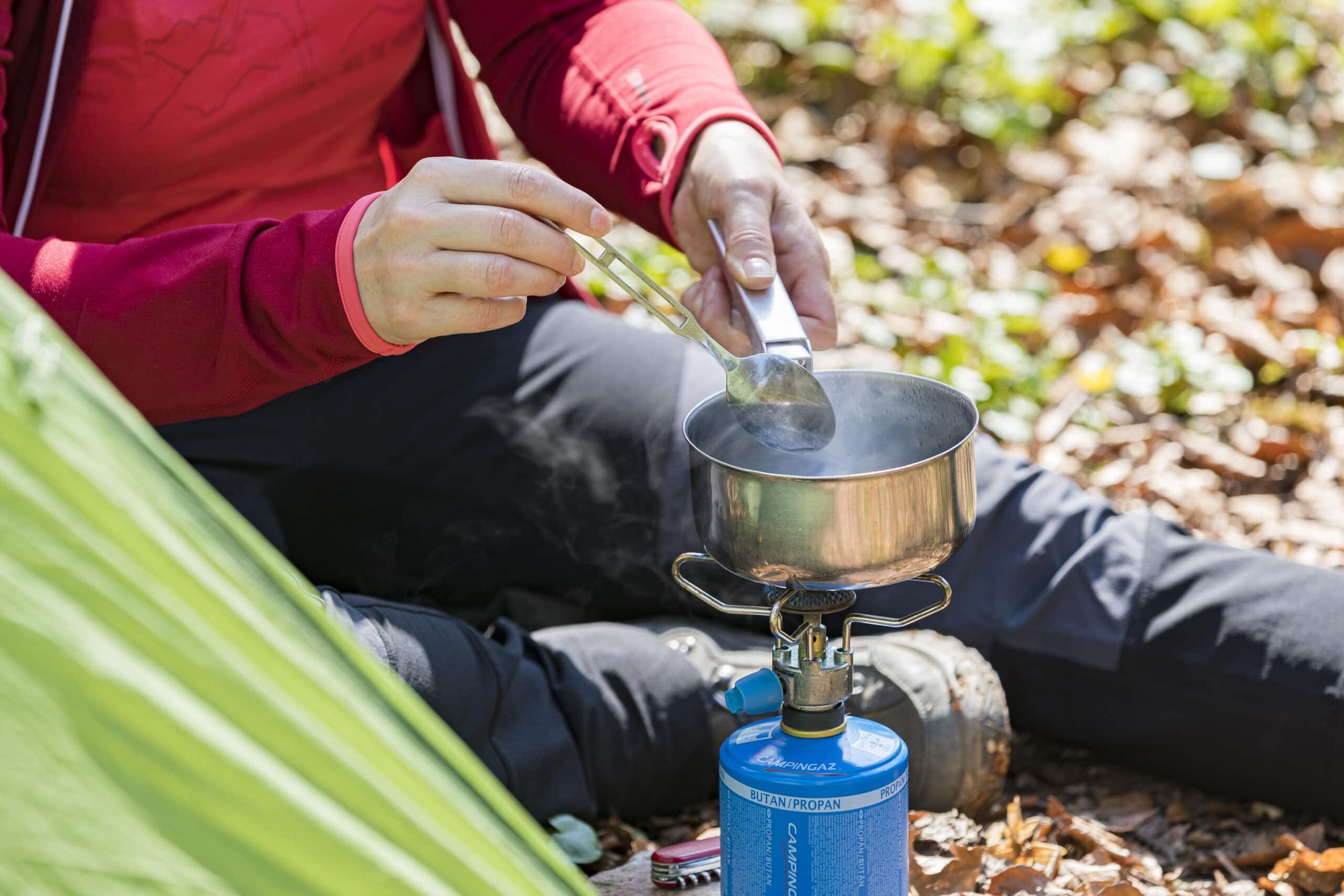 Person kocht mit Campingkocher und Gaskartusche im Wald, füllt Suppe mit Löffel in einen Topf, umgeben von Laub und Natur.