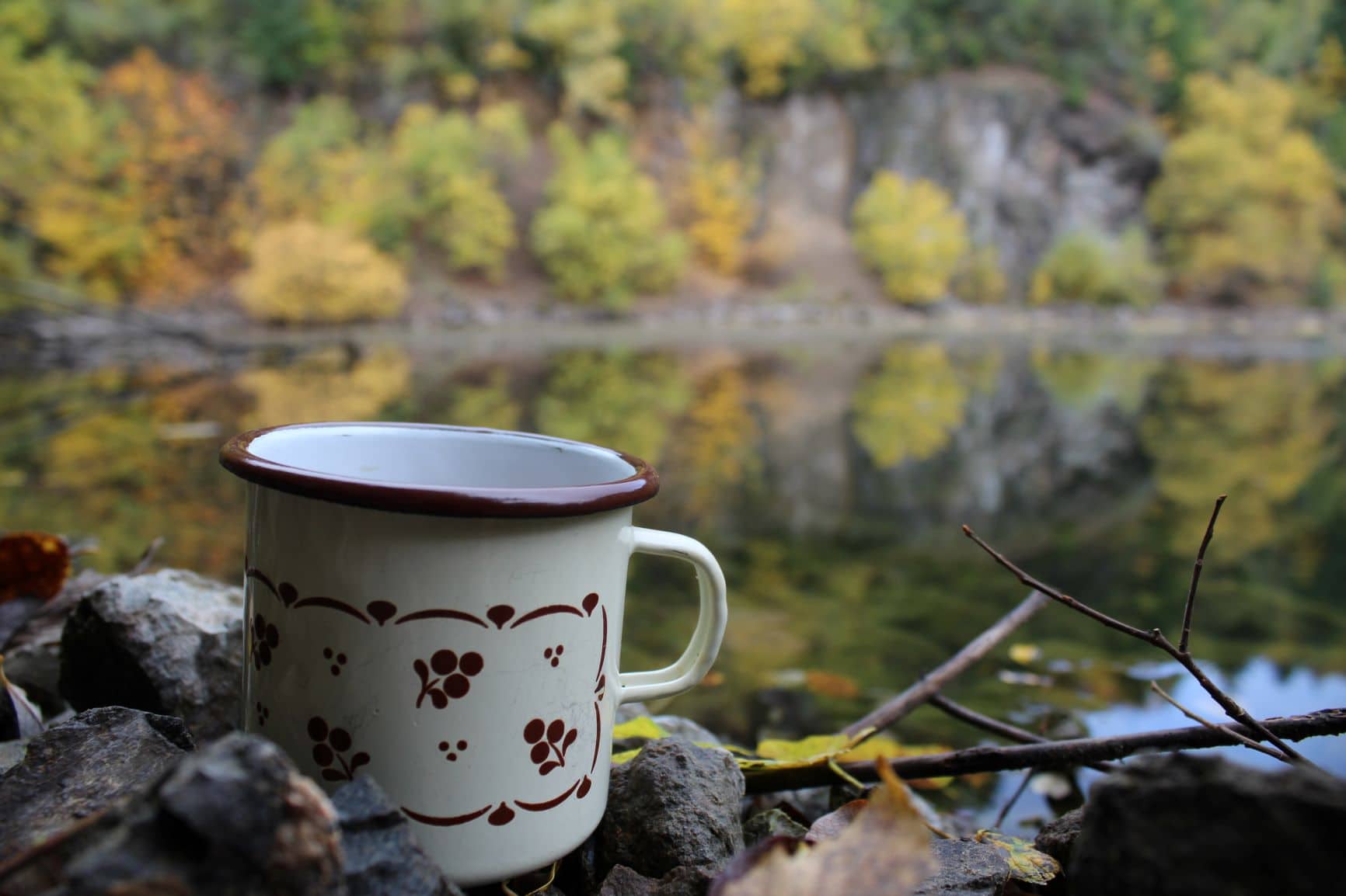 Robuste Edelstahltasse mit rotem Rand und Henkel steht auf Felsen in herbstlichem Wald mit gelb-grünem Laub im Naturpark Neckartal-Odenwald.
