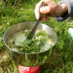 Person rührt mit einem Löffel eine warme Suppe in einem Camping-Kocher auf grüner Wiese im Freien, umgeben von Outdoor-Ausrüstung.