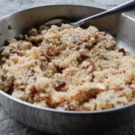Große Metallschüssel mit gekochtem Bulgur, vermischt mit getrockneten Aprikosen und Nüssen, Löffel liegt im Gericht.