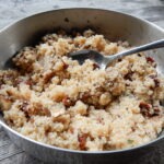 Metallschüssel mit gekochtem Couscous, vermischt mit getrockneten Früchten und Gewürzen, Löffel liegt im Gericht.