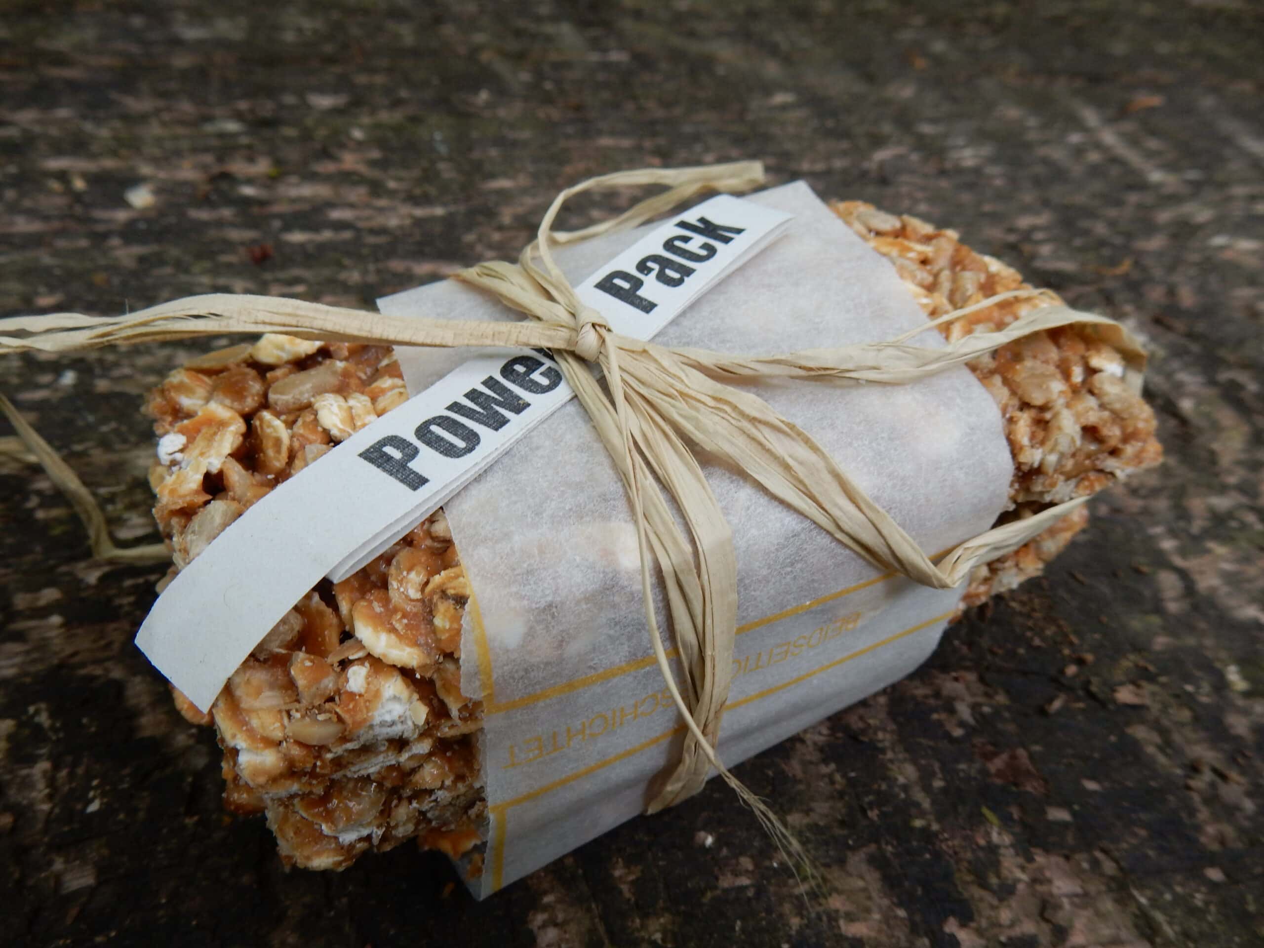 In Papier eingewickeltes Wanderproviant-Paket mit Nüssen und Snacks, fest mit Schnur gebunden, auf Waldboden.