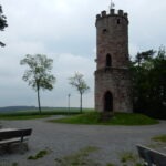 Steinerner Aussichtsturm mit Flagge im Naturpark Neckartal-Odenwald, umgeben von Wiesen und Bäumen, mit Sitzbänken und asphaltiertem Weg.