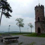 Historischer Aussichtsturm aus Buntsandstein im Naturpark Neckartal-Odenwald, umgeben von Wiesen und alten Bäumen, mit Sitzbänken und Wegweiser.