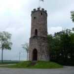 Historischer Aussichtsturm aus Naturstein mit Spitzdach und Flagge, umgeben von Wiese und Bäumen, typisch für ländliche Regionen im Neckartal-Odenwald.