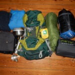 Ausgerüstet für Trekking: Rucksäcke, Schlafsack, Kochgeschirr, Erste-Hilfe-Set und Kleidung auf Holzboden ausgebreitet, bereit für eine Wanderung im Naturpark Neckartal-Odenwald.