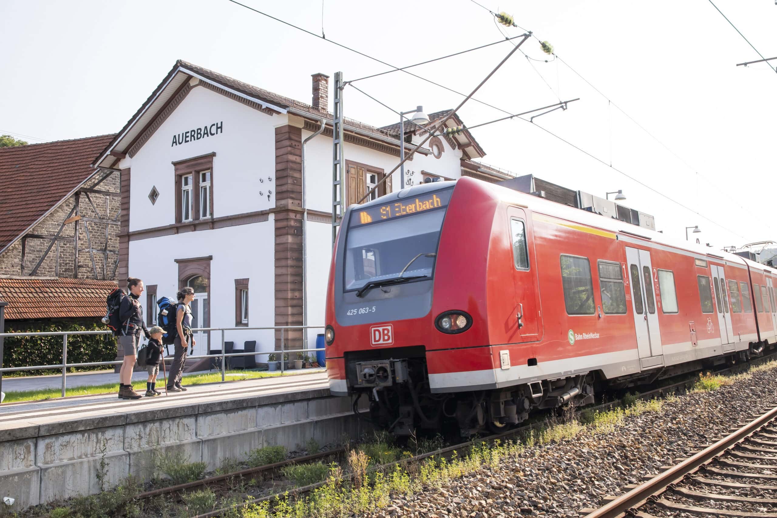 Drei Personen mit Rucksäcken steigen am Bahnhof Auerbach aus einem roten Regionalzug Richtung Odenwald aus, umgeben von historischem Bahnhofsgebäude.