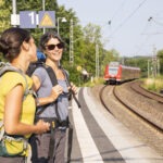 Zwei Personen mit großen Trekkingrucksäcken und Wanderausrüstung warten lächelnd auf Bahnsteig Gleis 1, während ein Regionalzug im Hintergrund vorbeifährt.