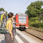Zwei Personen mit großen Trekkingrucksäcken stehen an Bahnsteigkante, warten auf Regionalzug Richtung Odenwald, umgeben von grünem Baumbestand und Fachwerkhäusern.