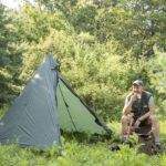 Person sitzt auf Baumstumpf im Wald, trinkt aus Becher und nutzt Campingkocher vor grünem Trekkingzelt im Naturpark Neckartal-Odenwald.