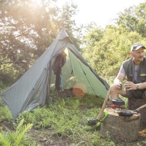 Mann sitzt entspannt auf Baumstumpf im Naturpark Neckartal-Odenwald, hält Metallbecher und nutzt Campingkocher vor grünem Trekkingzelt in sonnendurchflutetem Wald.