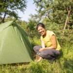 Person sitzt vor einem grünen Zelt auf einer sonnigen Wiese im Naturpark Neckartal-Odenwald, umgeben von dichtem Wald.