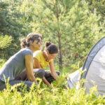 Zwei Personen richten im Naturpark Neckartal-Odenwald ein Zelt auf einer sonnigen Waldwiese mit dichtem Grünland auf.