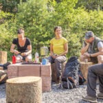 Vier Personen sitzen in einer Waldlichtung des Naturparks Neckartal-Odenwald auf Baumstammbänken und nutzen Campingausrüstung wie Thermoskannen und Besteck.