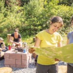 Zwei Personen im Naturpark Neckartal-Odenwald studieren gemeinsam eine Wanderkarte, während im Hintergrund andere an einer rustikalen Feuerstelle sitzen.