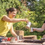 Zwei Personen gießen in einer Waldlichtung Getränke aus einer Thermoskanne in Metallbecher, umgeben von Campingausrüstung auf einem rustikalen Holztisch.