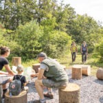 Drei Personen im Naturpark Neckartal-Odenwald besprechen eine Wanderkarte auf Baumstumpf-Sitzen an einer Raststelle mit Waldblick im Hintergrund.