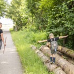 Zwei Personen mit Wanderausrüstung gehen auf einem asphaltierten Weg durch den grünen Wald im Naturpark Neckartal-Odenwald, begleitet von einem Kind mit ausgestreckten Armen.