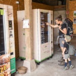 Zwei Personen mit Rucksäcken stehen vor einem automatischen Getränke- und Snackautomaten in einer rustikalen Hütte im Naturpark Neckartal-Odenwald. Regionale Produkte sind sichtbar.