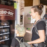 Zwei Personen mit Rucksäcken vor einer Regio-Box im Naturpark Neckartal-Odenwald, eine erwachsene Person wählt regionale Produkte aus.