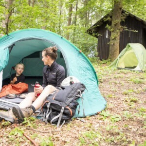 Zwei Personen sitzen vor einem grünen Zelt im schattigen Wald des Naturparks Neckartal-Odenwald, umgeben von Laub und Bäumen. Ein Rucksack und Snacks liegen daneben. Im Hintergrund weitere Zelte auf einer naturbelassenen Lichtung.
