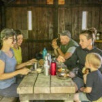Eine Gruppe von vier Personen sitzt in einer rustikalen Holzhütte im Naturpark Neckartal-Odenwald und isst gemeinsam an einem langen Holztisch mit einfachen Mahlzeiten, Trinkflaschen und Snacks.