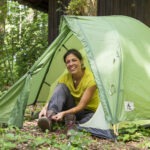 Person sitzt lächelnd im grünen Trekkingzelt im schattigen Wald des Naturparks Neckartal-Odenwald und bindet Wanderschuhe.