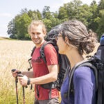 Zwei Wanderer mit großen Rucksäcken und Trekkingstöcken laufen durch ein hochstämmiges Getreidefeld mit Wald im Hintergrund im Naturpark.