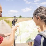 Zwei Personen mit Wanderausrüstung und Trekkingstöcken besprechen eine Wanderkarte auf einem Feldweg in offener Landschaft mit blauem Himmel.