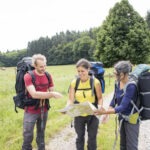 Drei Wanderer mit großen Rucksäcken und Wanderstöcken besprechen gemeinsam eine Karte auf einem Feldweg im Naturpark Neckartal-Odenwald, umgeben von Wiesen und Wald.