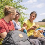 Drei Personen in lockerer Kleidung sitzen an einem Feldweg im Naturpark, essen gemeinsam Brot und unterhalten sich, umgeben von grünem Gestrüpp und einer offenen Landschaft.
