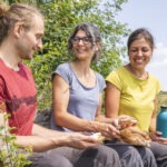 Drei Personen sitzen auf einer Holzbank in einer grünen Landschaft und essen gemeinsam Brot. Im Hintergrund ist eine offene Wiesenlandschaft mit Hecken und einem Wanderweg zu sehen.