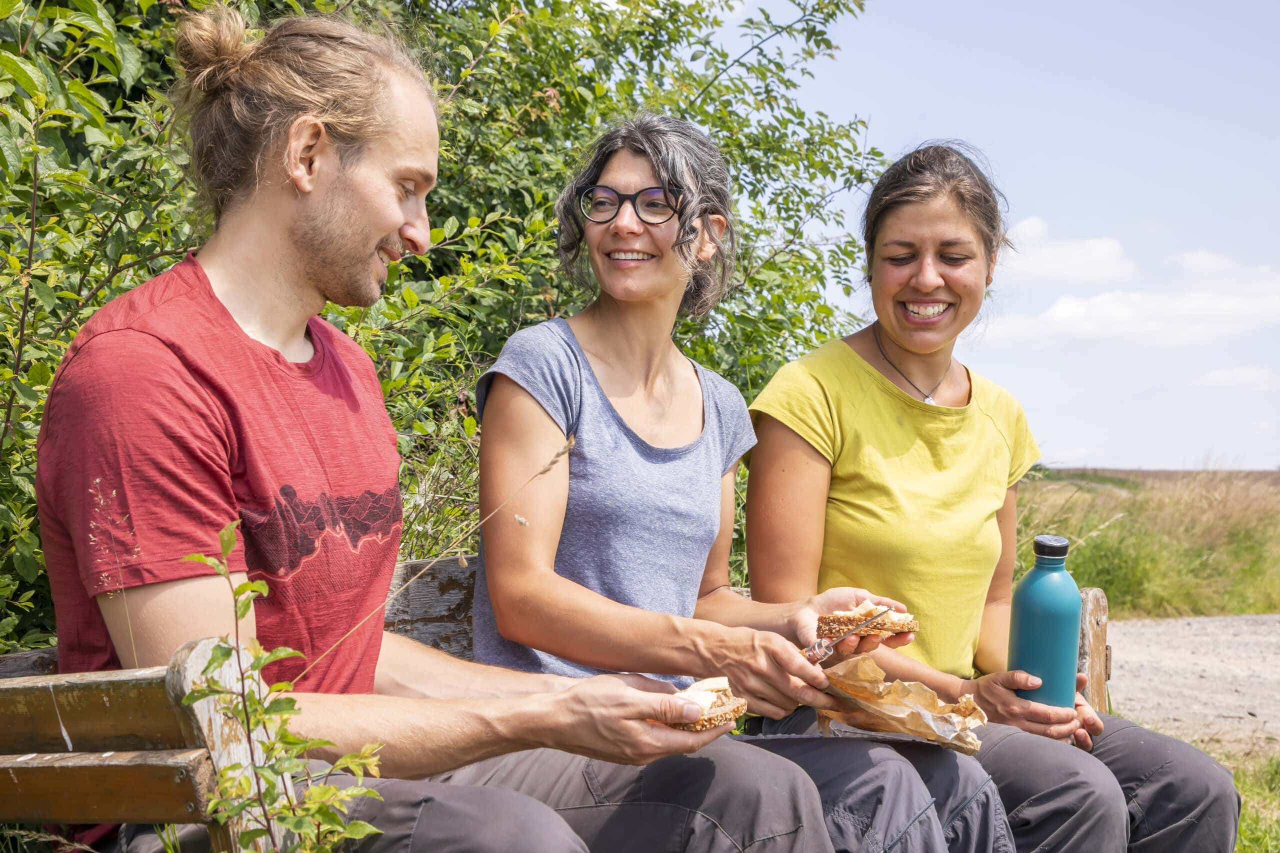 Drei Personen sitzen auf einer Holzbank in einer grünen Landschaft und essen gemeinsam Brot. Im Hintergrund ist eine offene Wiesenlandschaft mit Hecken und einem Wanderweg zu sehen.